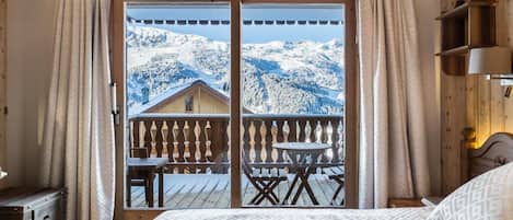 A bedroom with a large bed, wooden headboard, and a view of snow-covered mountains through a balcony.