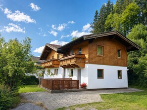 A two-story wooden house with a balcony, surrounded by greenery and trees.