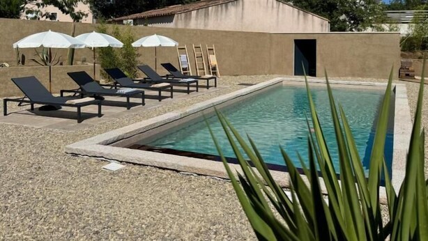 Une piscine rectangulaire entourée d’une zone de gravier et d’un bâtiment doté d’un toit en tuiles, avec des chaises longues et des parasols à proximité.