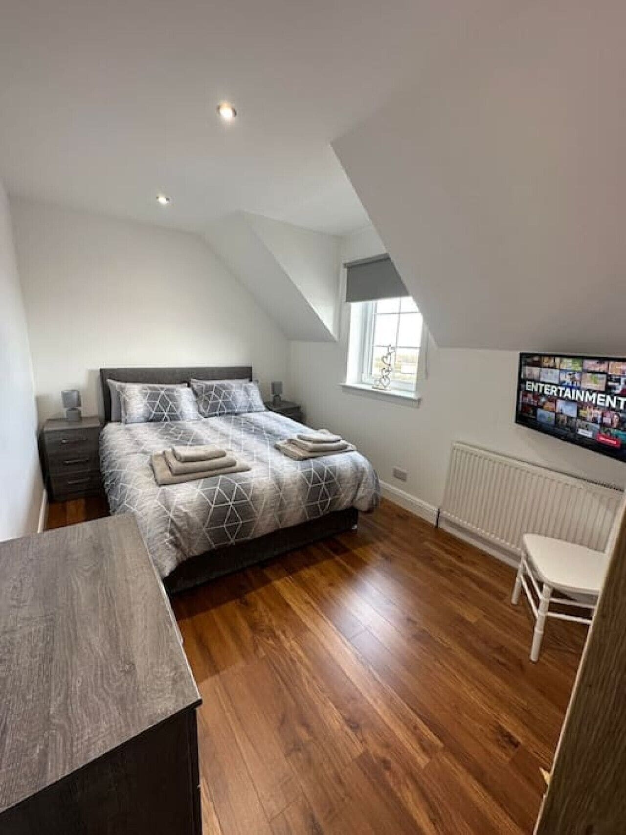 A bedroom with a bed, a desk, a chair, a window, and a wall-mounted TV.