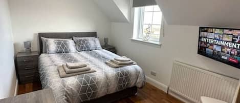 A bedroom with a bed, a desk, a chair, a window, and a wall-mounted TV.
