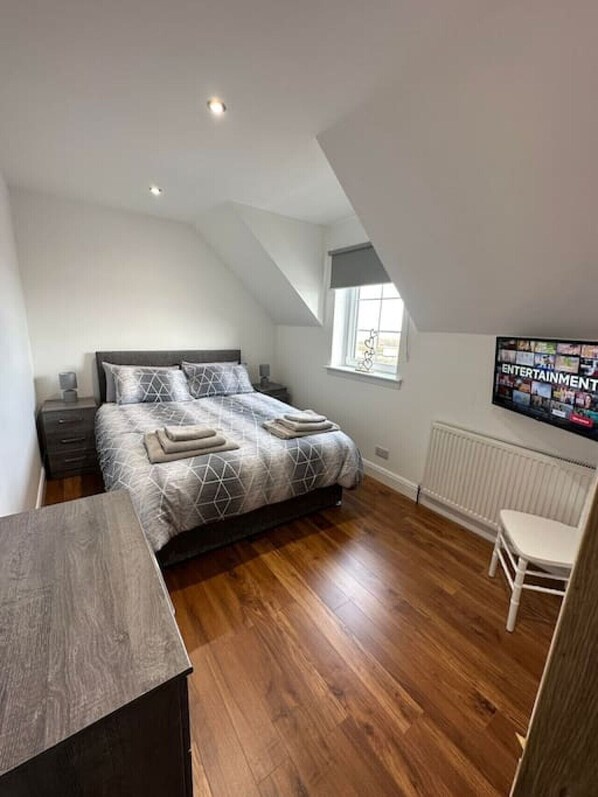 A bedroom with a bed, a desk, a chair, a window, and a wall-mounted TV.