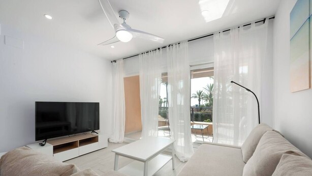 A modern living room with a flat-screen TV, a sofa, a coffee table, and a ceiling fan. - Sotogrande Beach Apartment (San Roque)