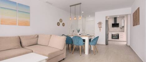 Una sala de estar moderna con un sofá y una mesa de comedor con sillas. Se observa una zona de cocina con electrodomésticos.