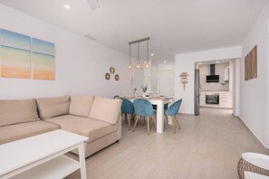 Una sala de estar moderna con un sofá y una mesa de comedor con sillas. Se observa una zona de cocina con electrodomésticos.