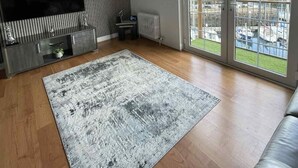 A modern living room with a large TV, a sofa, a patterned rug, and a view of a marina through large windows. - Seaview Apartment in Banff (Aberdeenshire)