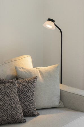 Een bank met twee kussens, een effen en een met dessin, tegen een egale muur met een vloerlamp.