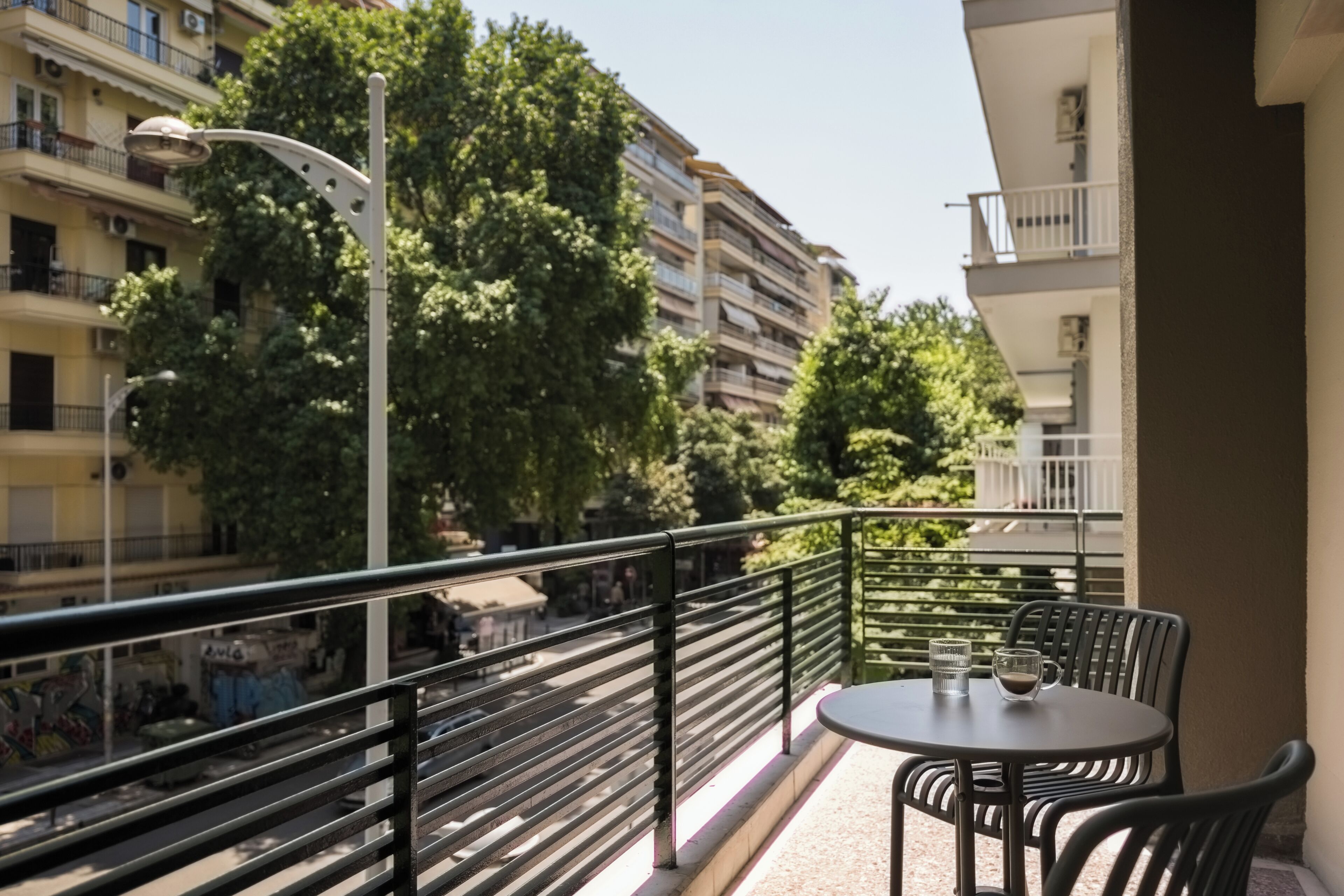 A balcony with a table and chairs, overlooking a street with buildings and trees.