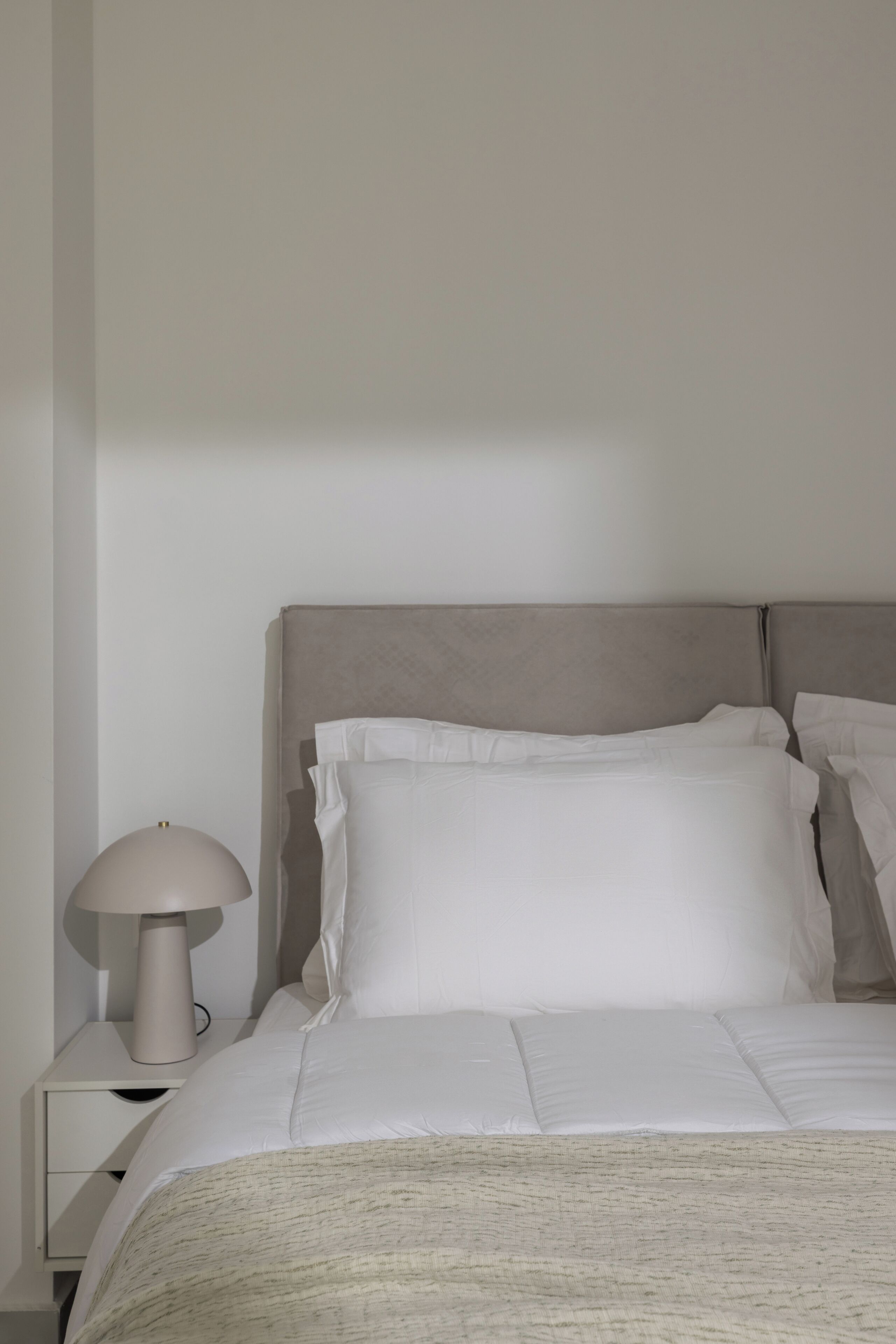 Ein ordentlich bezogenes Bett mit weißen Leinen und Kissen, eine Nachttischlampe und eine schlichte Wand.