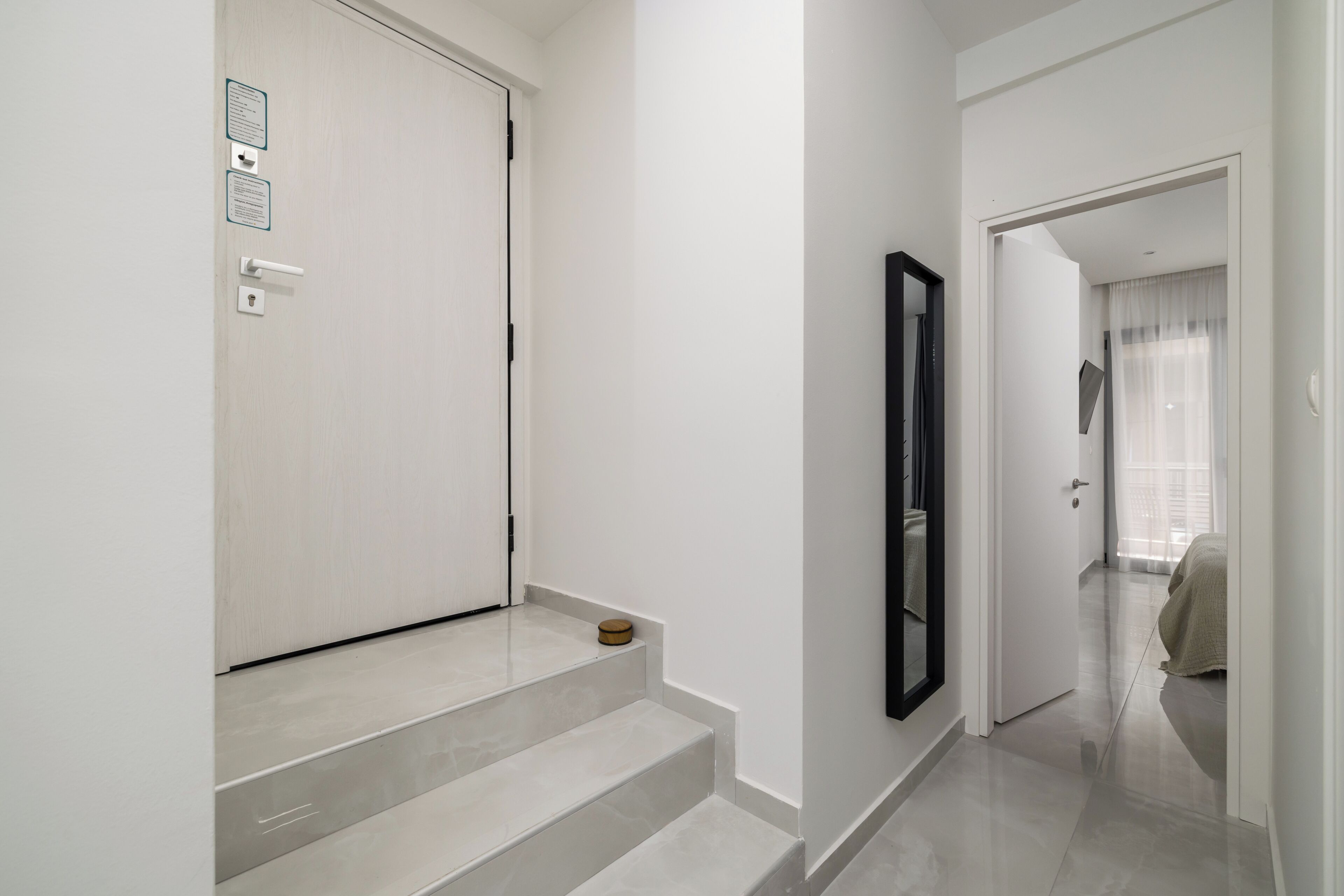 Een moderne hal met een witte deur, een klein houten krukje en een spiegel aan de muur.