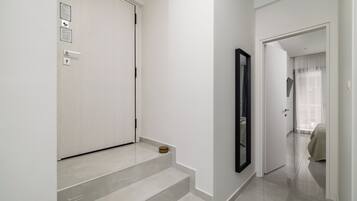 Een moderne hal met een witte deur, een klein houten krukje en een spiegel aan de muur.