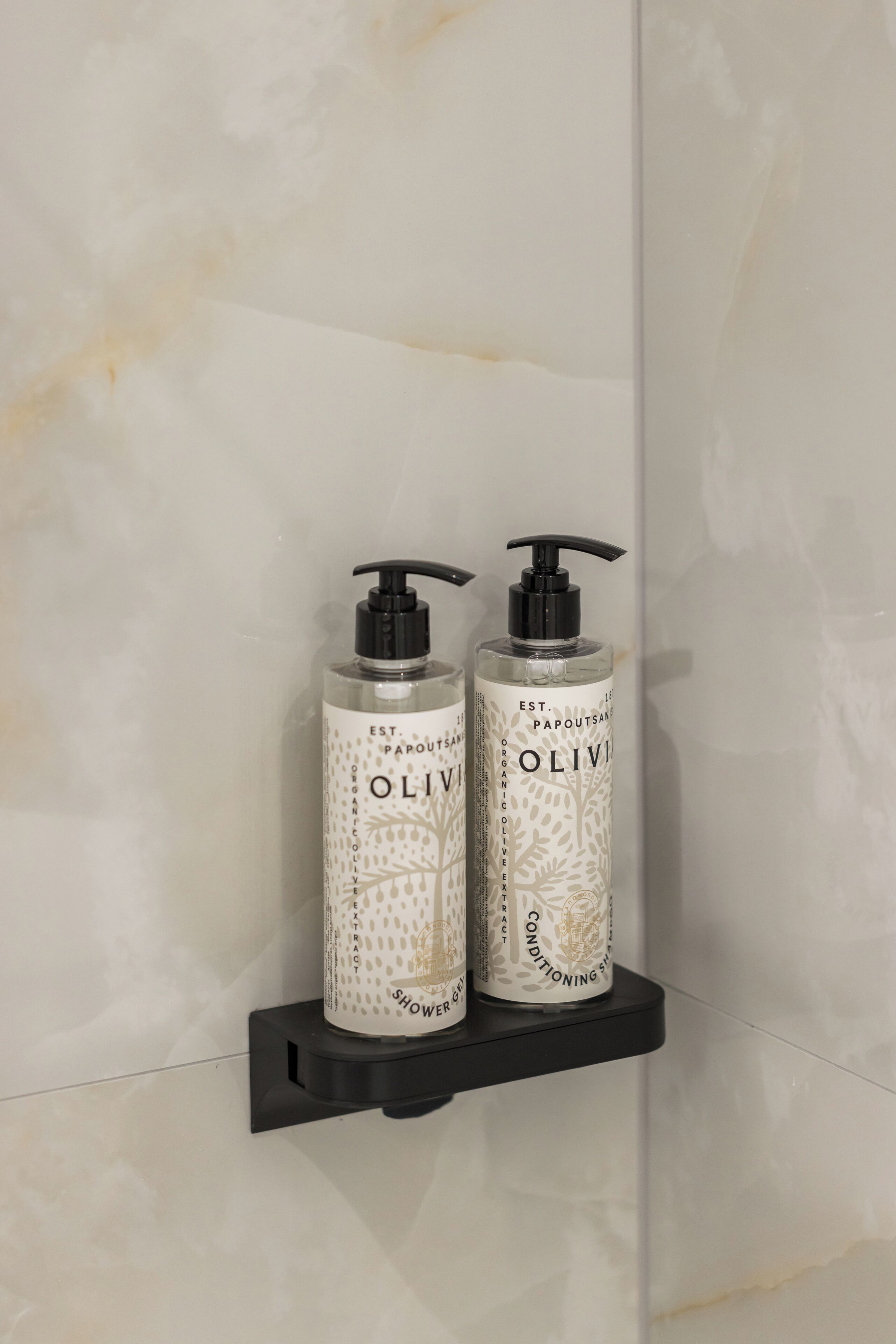 Deux bouteilles de gel douche à base d’extrait d’olive biologique sont posées sur une étagère noire, adossée à un mur en marbre.