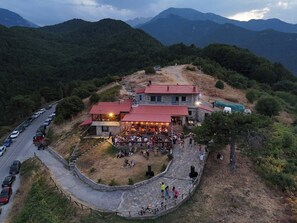 A building with a red roof, surrounded by a stone pathway, and a parking area with cars. - Olympus Refuge D (Dio-Olympos)