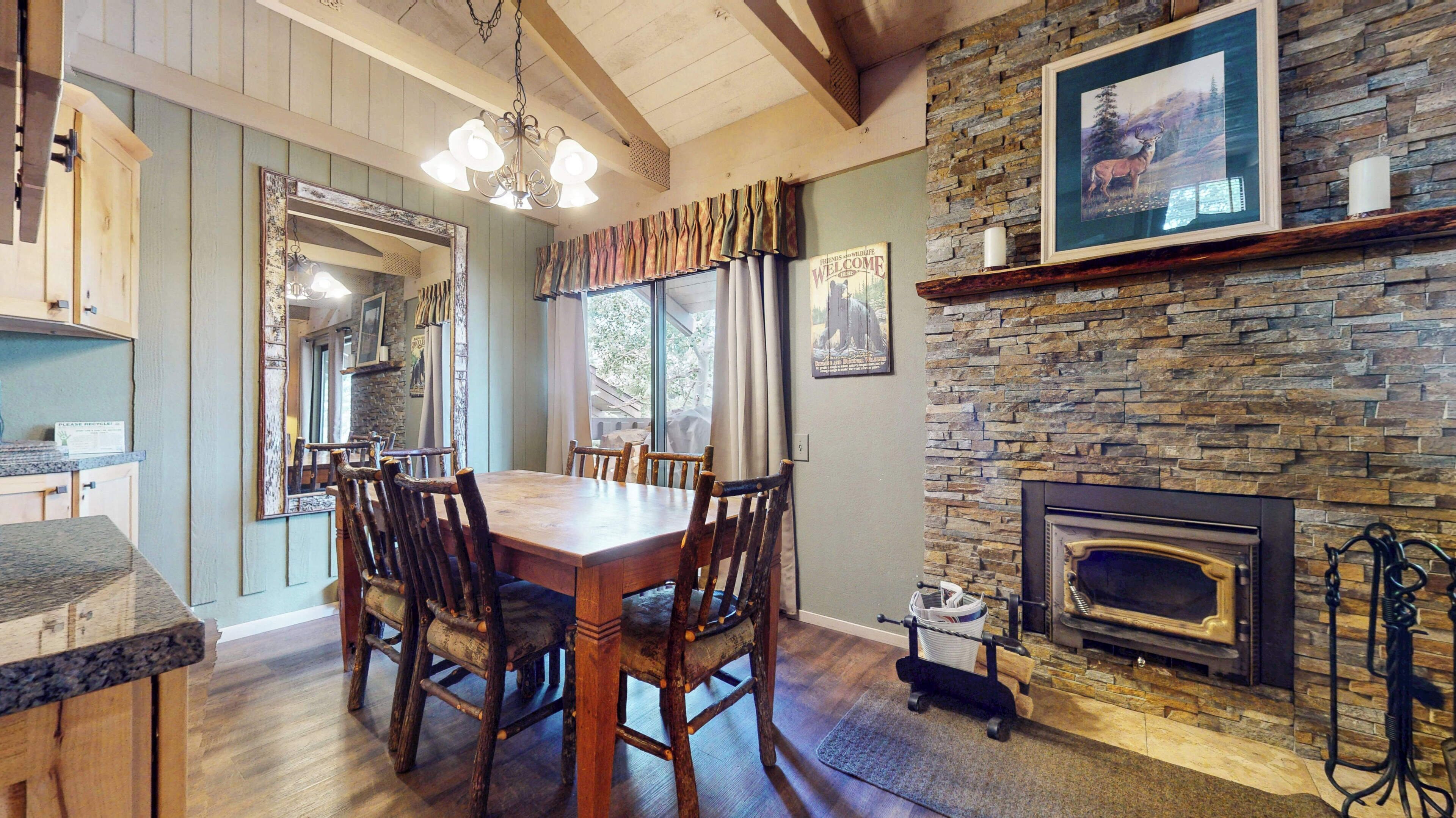 A dining area with a wooden table and chairs, a stone fireplace, and a view of the outdoors through large windows.