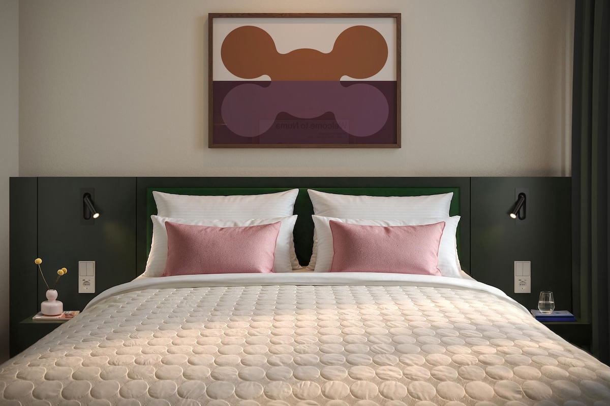 A bed with a quilted cover, two pillows, a green headboard, and a wall-mounted abstract painting.