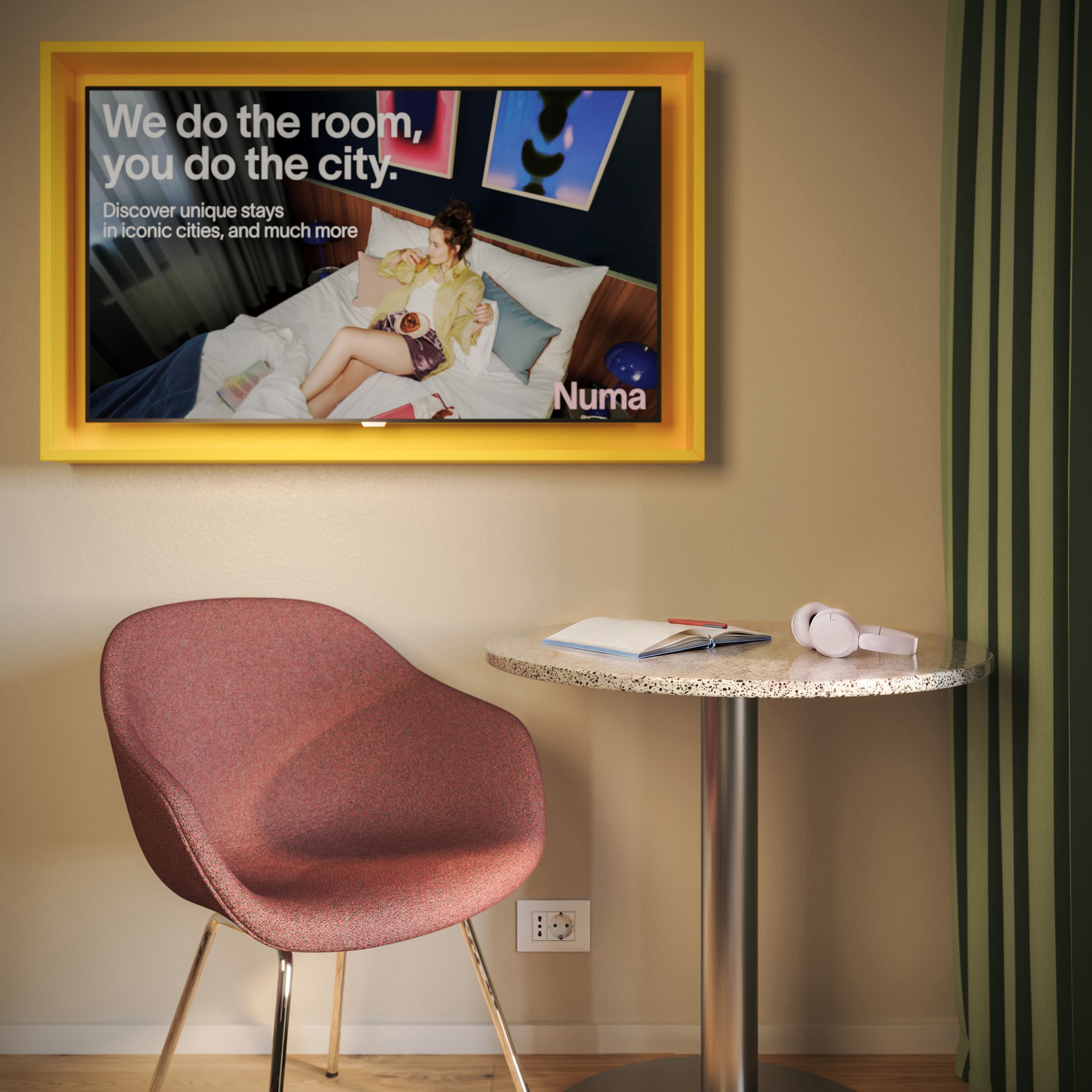Una habitación de hotel con un anuncio enmarcado en la pared, una mesa redonda con un cuaderno, bolígrafo y auriculares, y una silla roja.
