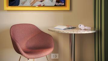 Una habitación de hotel con un anuncio enmarcado en la pared, una mesa redonda con un cuaderno, bolígrafo y auriculares, y una silla roja.