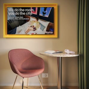 A hotel room with a framed advertisement on the wall, a round table with a notebook, pen, and headphones, and a red chair. - Numa Milan Manfredo (Milan)