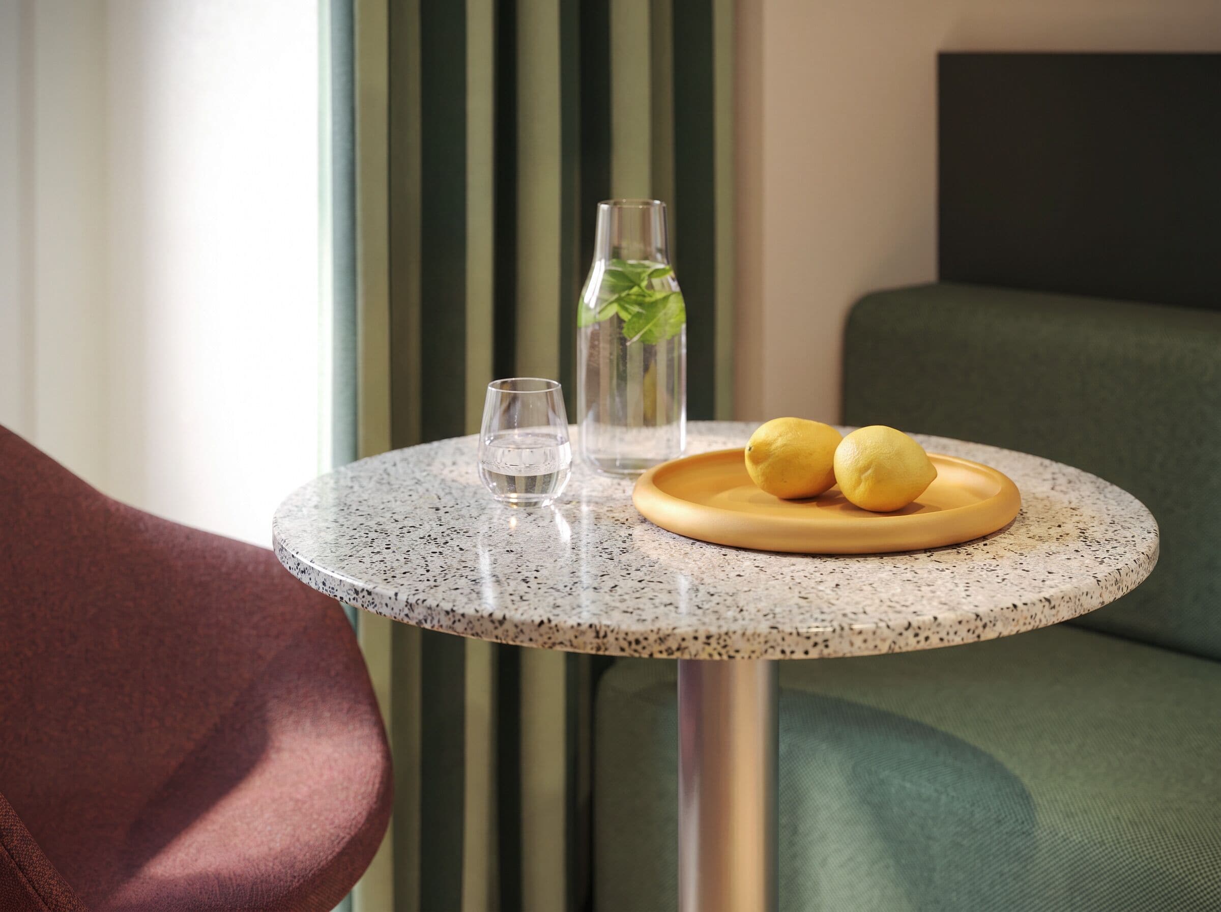 Una mesa redonda con superficie moteada, una botella de vidrio transparente con una planta verde, dos limones en un plato de madera y un vaso de agua.