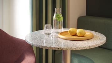Una mesa redonda con superficie moteada, una botella de vidrio transparente con una planta verde, dos limones en un plato de madera y un vaso de agua.