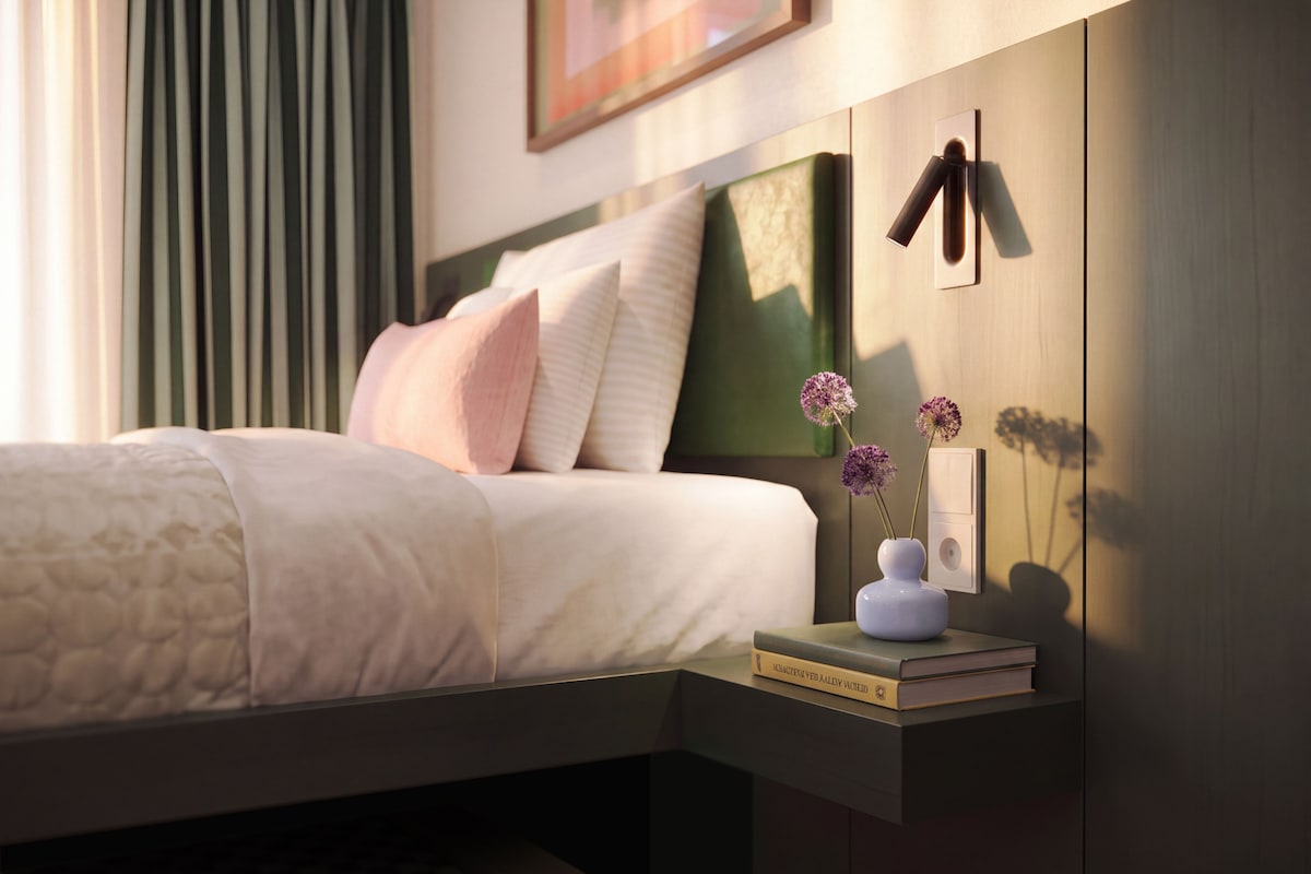 A neatly made bed with pillows, a bedside table with a book and a vase of flowers, and a wall-mounted light switch.