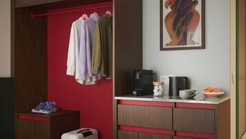 Quarto com parede vermelha, um armário de madeira, cabideiro com várias peças de roupa, cafeteira, caneca e uma tigela de frutas.