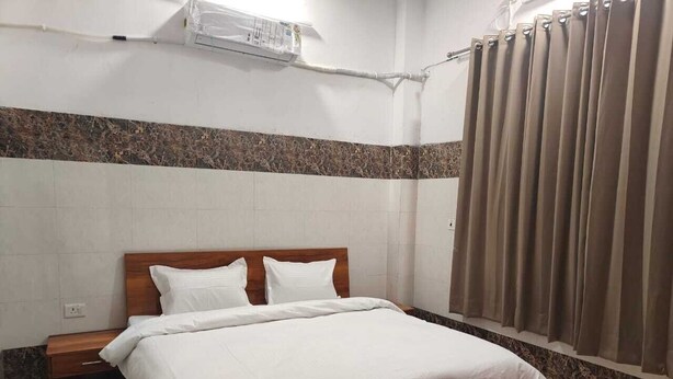 A hotel room with a wooden bed, white bedding, a nightstand, a wall-mounted air conditioner, and a window with curtains. - The Hotel Junction Palace  (Aligarh)