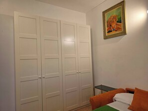 Ein Schlafzimmer mit großem weißen Kleiderschrank, einem kleinen Tisch, einem Bett mit orange-weißer Bettwäsche und einem gerahmten Gemälde an der Wand.