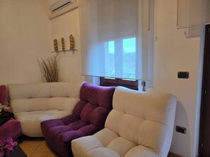 A corner living room with a beige sectional sofa, a purple accent chair, a small plant, and a window with blinds. - Maison Galliano  Verona (Verona)