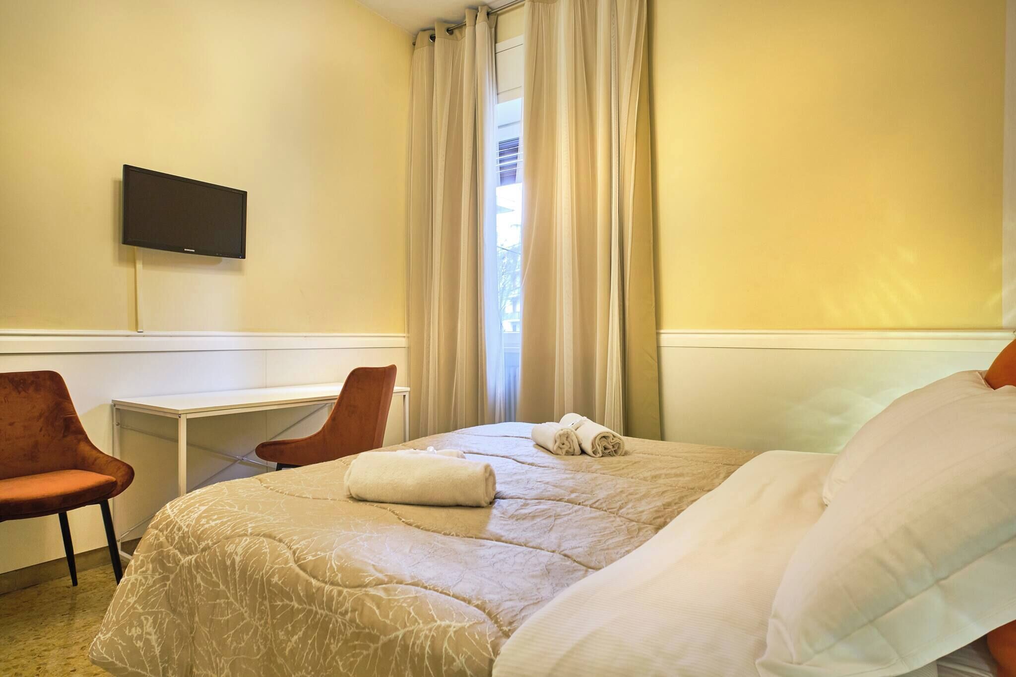 A hotel room with a bed, a desk, a chair, and a television on the wall.