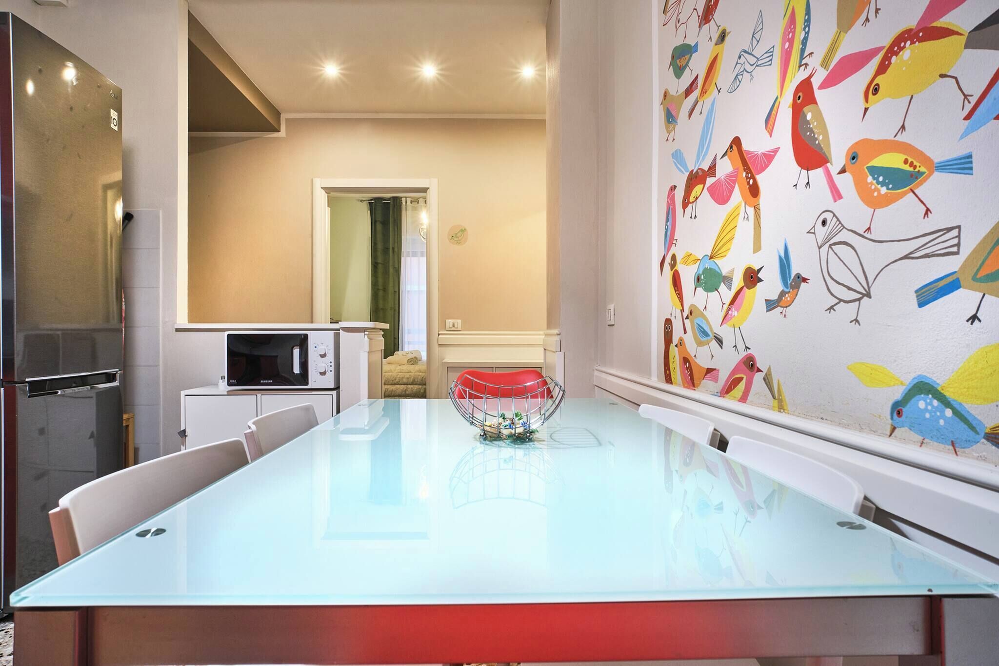 Un espace repas moderne avec une longue table, des chaises, un four à micro-ondes et un mur décoré d’illustrations colorées d’oiseaux.
