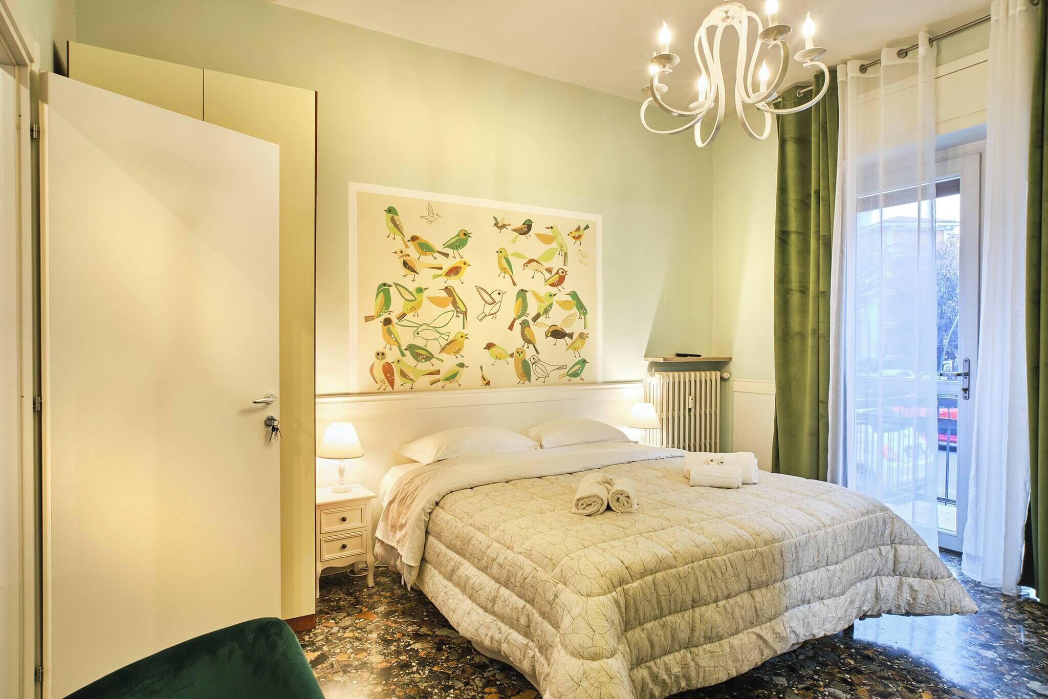 A hotel room with a bed, a nightstand, a wall-mounted artwork of birds, a radiator, and a view through a window with curtains.
