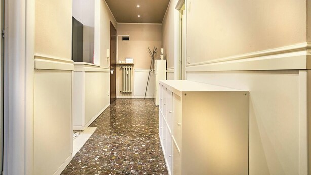 Um corredor estreito com piso de pedra polida, paredes brancas e um radiador.