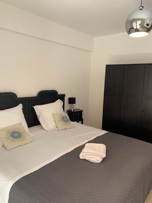 Una cama bien hecha con edredón blanco, colcha acolchada gris y una toalla doblada. Junto a la cama hay una mesita de noche negra con una lámpara y un adorno. Se ve un armario oscuro al fondo.