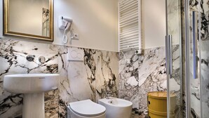 Een badkamer met een marmeren wand, een wastafel op voetstuk, een toilet, een bidet en een afvalbak in goudkleur.