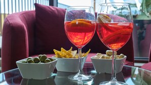 Twee glazen rode drank met oranjeslices, een schaal groene olijven en een schaal tortilla chips op een glazen tafel.