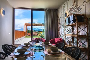 A dining area with a table set for four, a red chair, a bamboo shelf with glassware, and a view of mountains through a large window. - Maison Bra 10 - Purple (Verona)