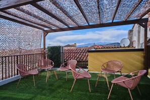 A covered outdoor seating area with artificial grass, metal chairs, and a view of buildings and mountains in the background. - MAISON BRA RED Verona (Verona)