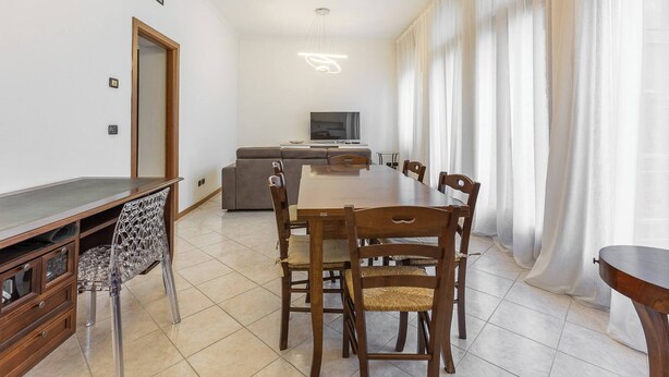 A dining area with a long wooden table and chairs, a sofa, a television, and a large window with sheer curtains. - Stanza con bagno privato 9 - uso cucina Bastioni (Verona)