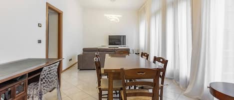 A dining area with a long wooden table and chairs, a sofa, a television, and a large window with sheer curtains.