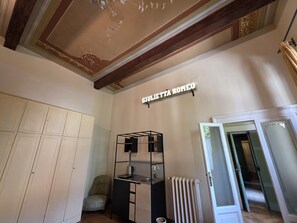 Ein Zimmer mit einer dekorativ verzierten Decke, einem Schild mit der Aufschrift 'CIVILETTA ROMEO', einem Heizkörper, einem kleinen Schreibtisch mit Wasserkocher und einem Stuhl.