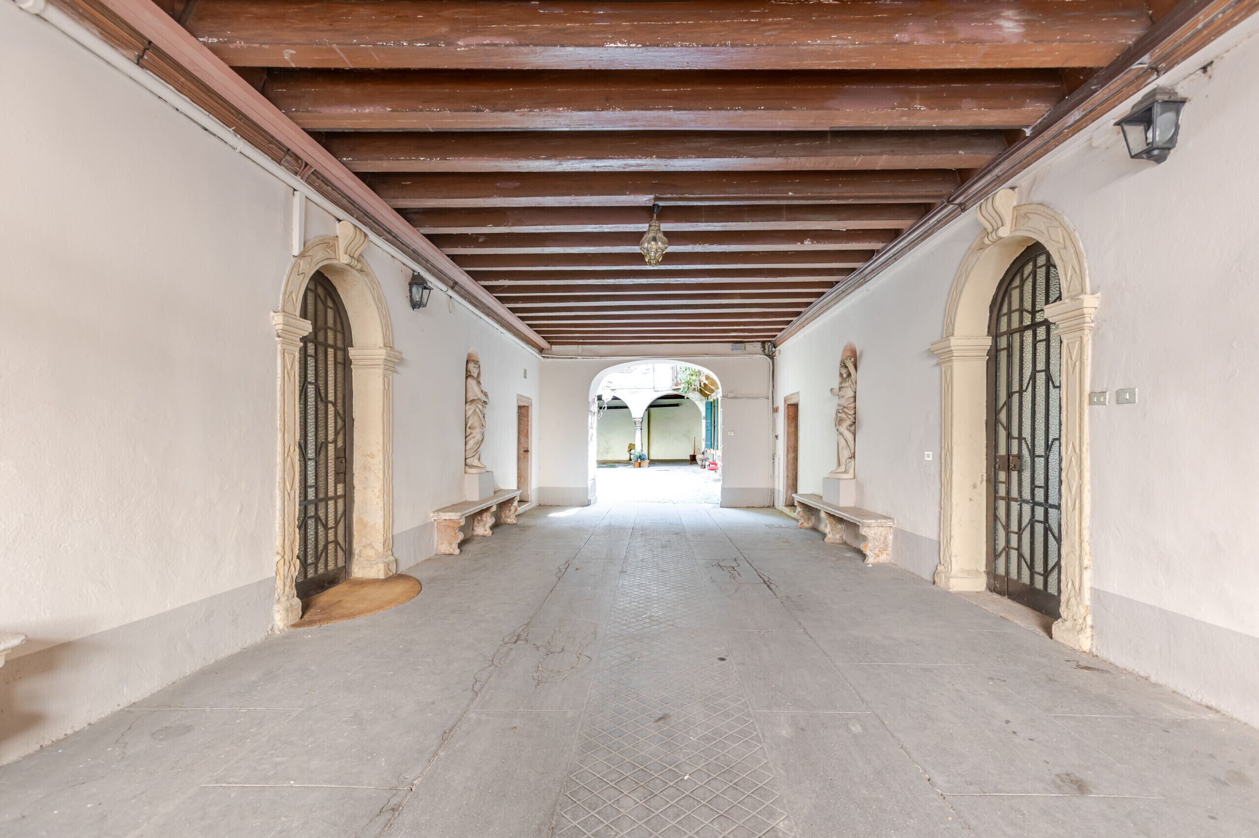 A corridor with a tiled floor, wooden beams on the ceiling, and arched doorways on both sides.