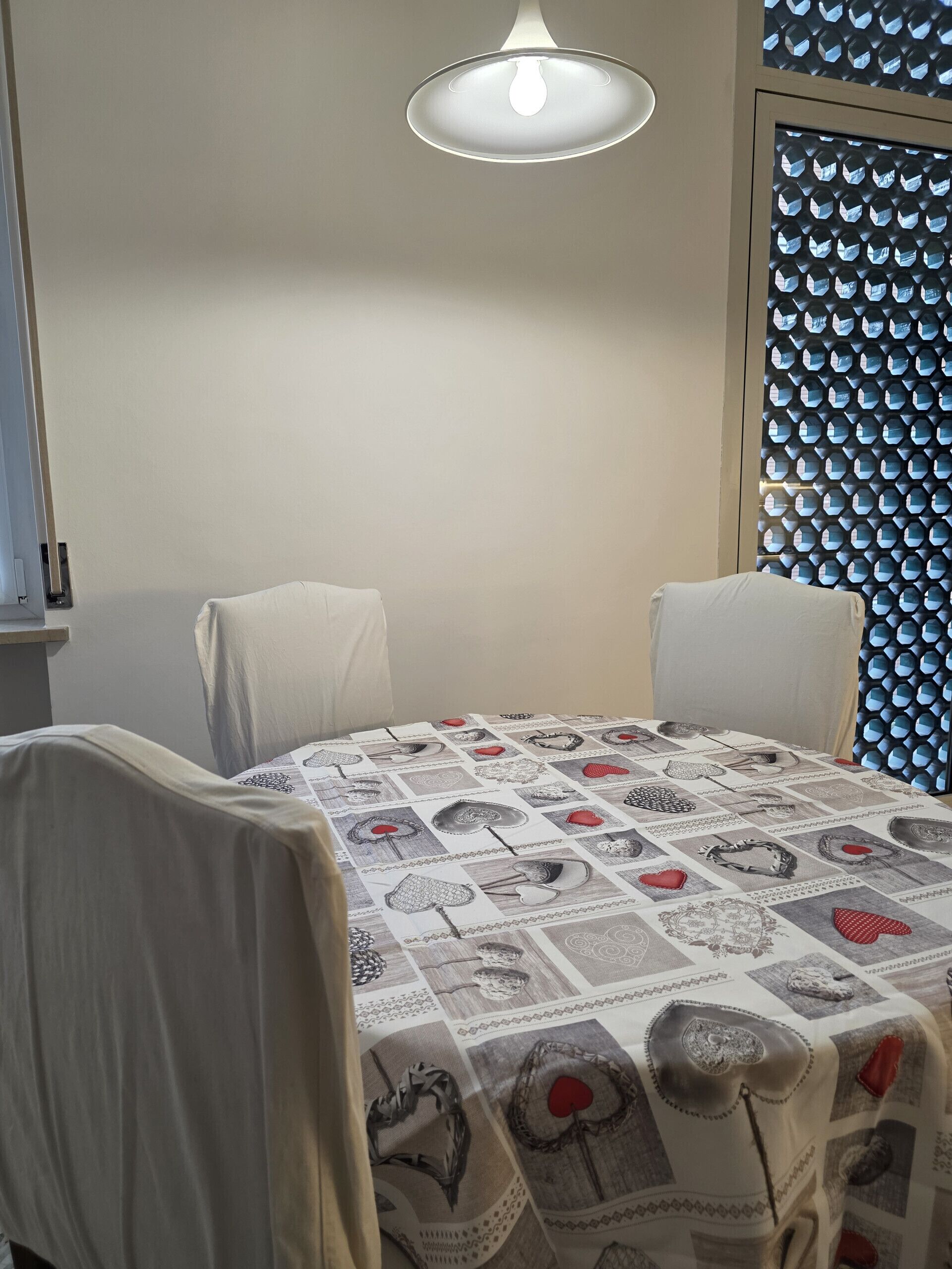 A dining area with a table covered in a patterned tablecloth, two chairs, a hanging light fixture, and a wall with a geometric pattern.
