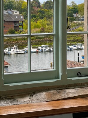 Blick durch ein Fenster auf einen Yachthafen mit vertäuten Booten, ein Gebäude mit einem Balkon und Grün im Hintergrund.