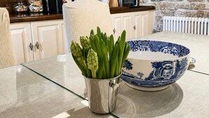 A kitchen with white cabinets, a stone wall, and a glass-top table with a blue and white bowl and a metal pitcher with green vegetables. - Central Boutique 2 Bed Cottage – Sleeps 6 (Lymington)
