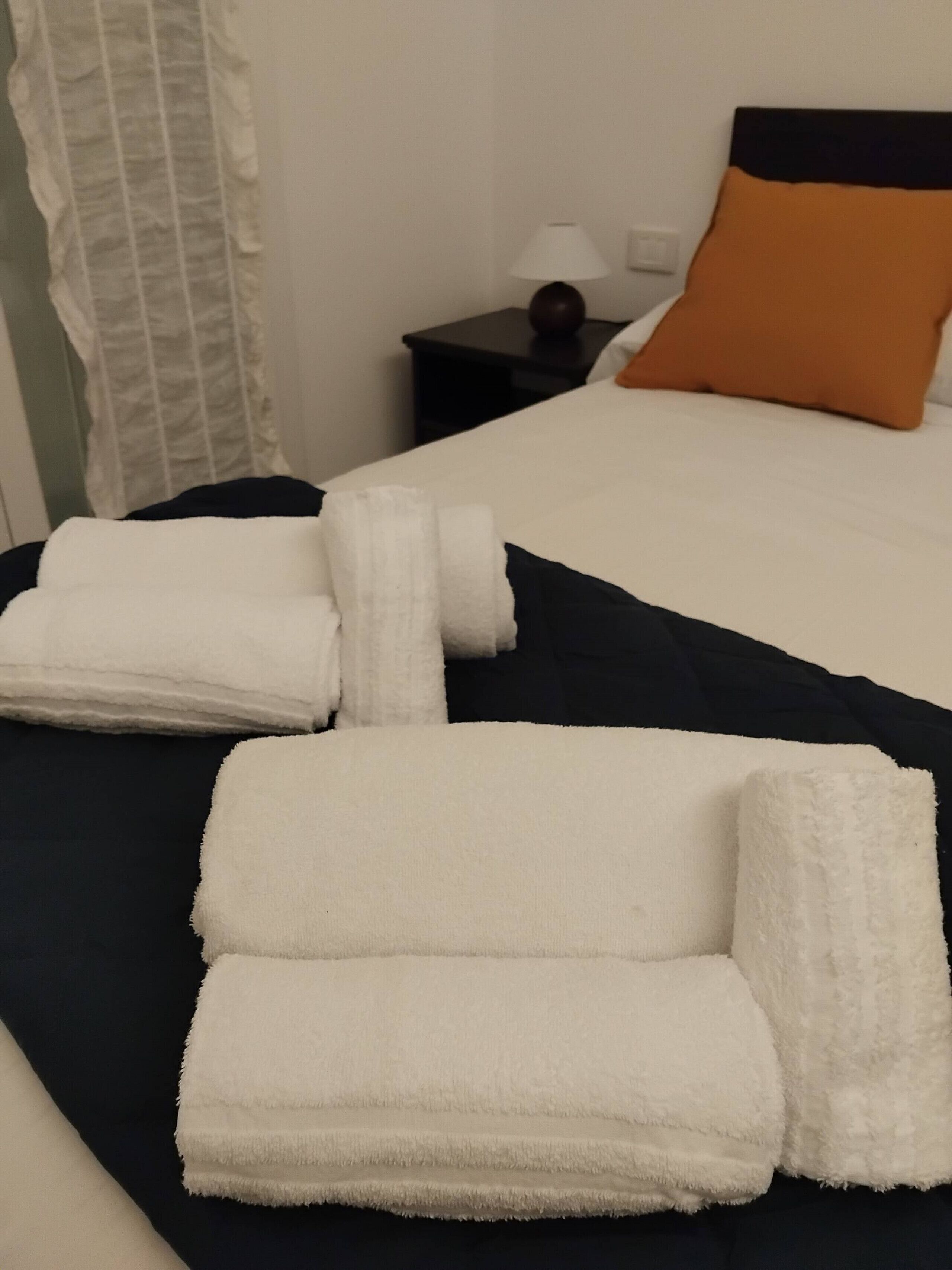 A neatly made bed with white linens and a black bedspread, a nightstand with a lamp, and a pillow with an orange accent.