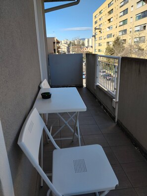 Ein Balkon mit einem weißen Tisch und Stühlen, einer kleinen schwarzen Schale und Blick auf eine Stadtlandschaft mit Gebäuden und parkenden Autos.