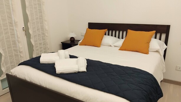 A neatly made bed with a dark headboard, white and blue bedding, and two orange pillows. - Appartamento La Sosta (Verona)