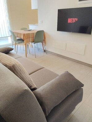 Ein Wohnzimmer mit einer Couch, einem Esstisch mit Stühlen und einem an der Wand befestigten Fernseher, der 'NETT' anzeigt.
