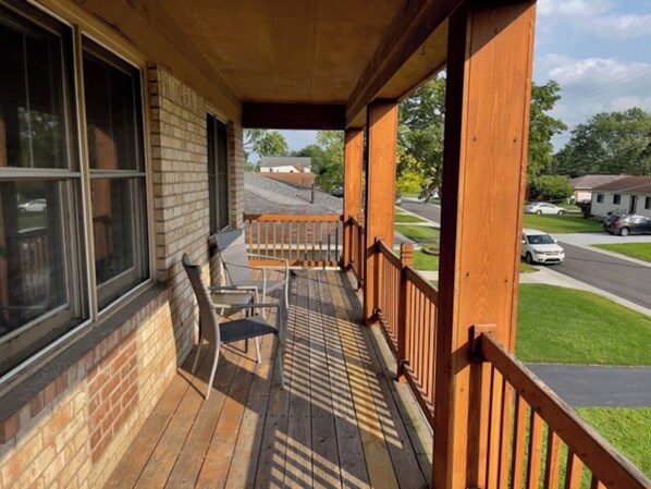 Una terraza de madera con barandilla, un muro de ladrillo y vista a una calle con casas.